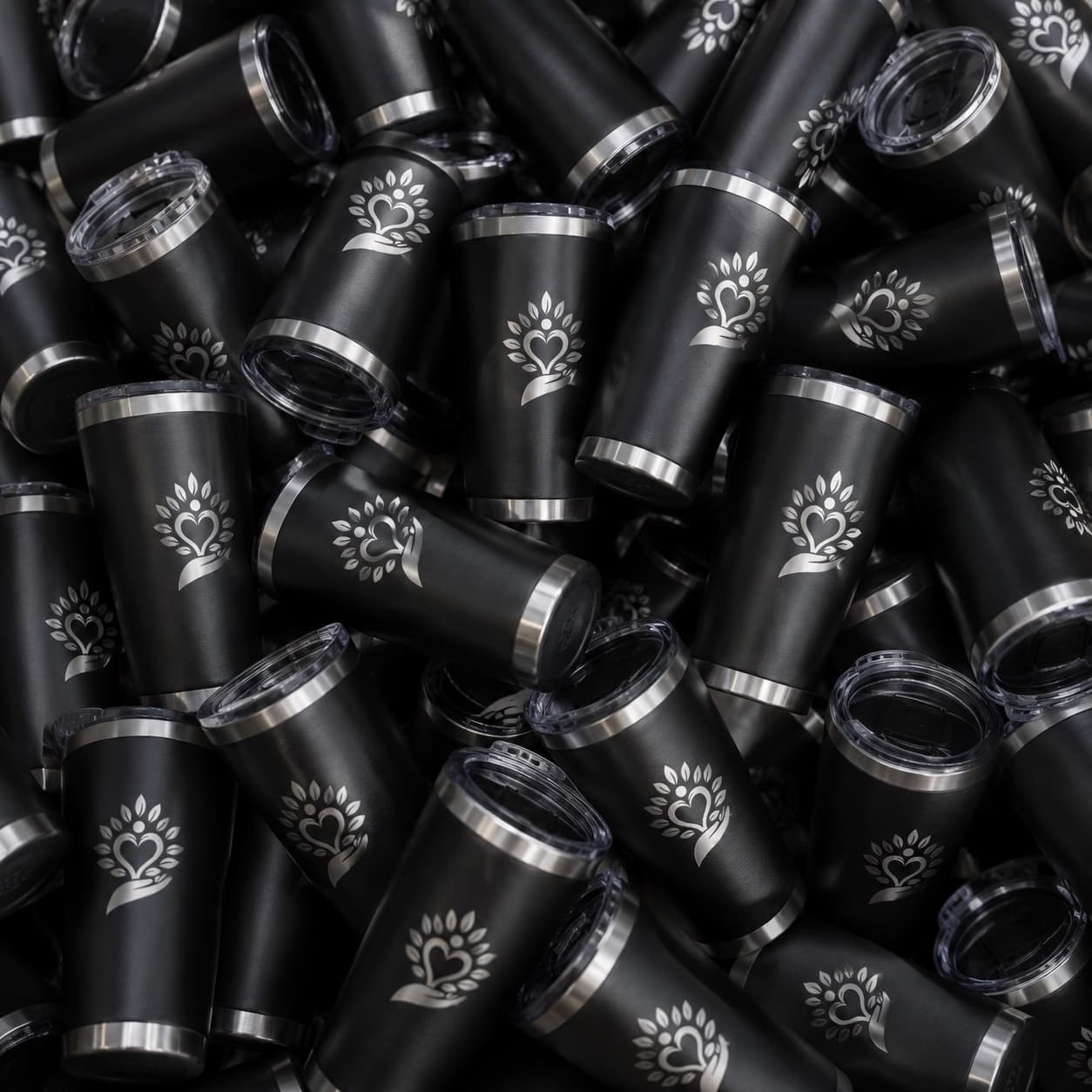 A pile of branded matte-black insulated tumblers, each engraved with a silver tree and heart company logo